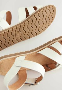 Sandales blanches à lanières avec une semelle texturée beige. La partie supérieure présente des lanières en cuir lisse et une boucle pour ajustement.