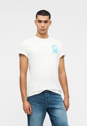 Νεαρός άνδρας που φοράει λευκό μπλουζάκι με μανίκια γυρισμένα και μπλε σχέδιο στο στήθος και μπλε τζιν, στέκεται μπροστά σε απλό φόντο.