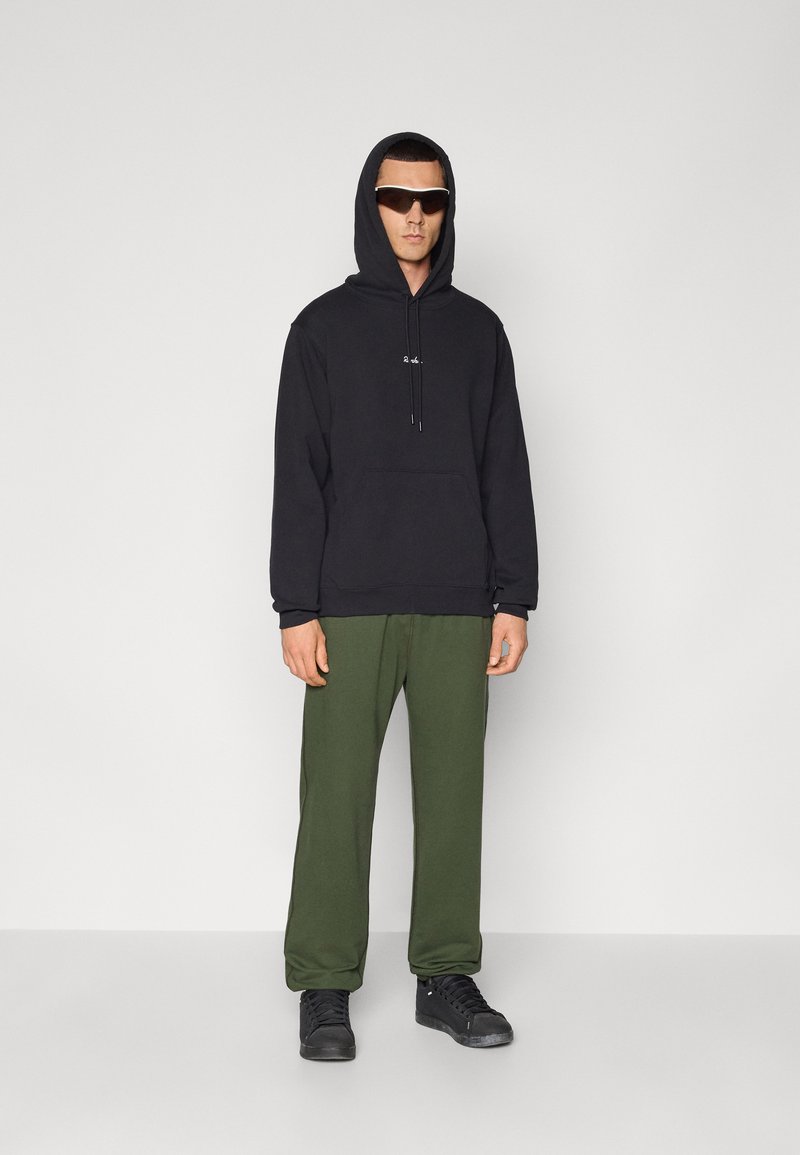 Sort hoodie med en frontlomme, grøn sweatpants og sorte sneakers. Model iført mørke solbriller, står mod en neutral baggrund.