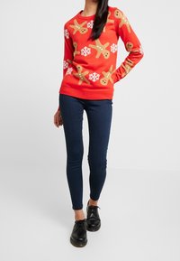 Pull rouge avec motifs de bonhomme en pain d'épice et flocons de neige, col rond, poignets côtelés, associé à un jean skinny bleu foncé et des chaussures noires.