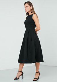 Black sleeveless dress with a fitted bodice, pleated skirt, and knee-length hem. Pair of black strappy high-heeled sandals.