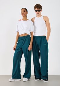 Türkise Sporthose mit grünen Seitestreifen und elastischem Bund, kombiniert mit einem weißen Crop-Top und einem weißen Tank-Top.