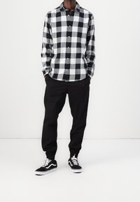 Camicia a quadri bianca e nera con bottoni, pantaloni neri e sneaker nere con dettagli bianchi. La camicia presenta un'orlo arrotondato e maniche lunghe.