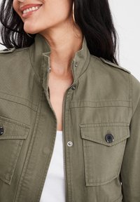 Giacca utility verde oliva con zip frontale, tasche sul petto con bottoni e colletto alla francese. Il tessuto sembra resistente con una finitura texture.