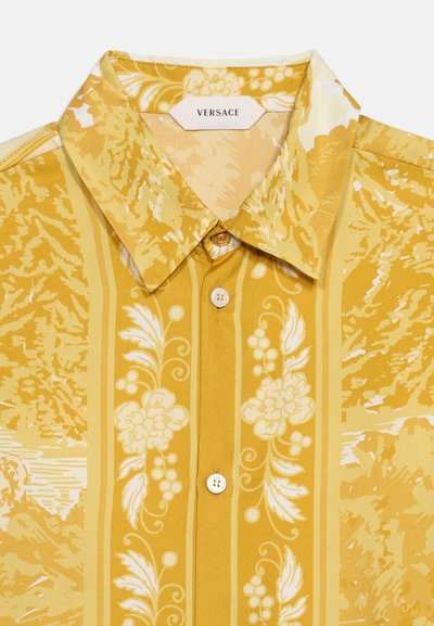 Κίτρινο πουκάμισο Versace με μυτερό γιακά, κατακόρυφα ανθικά και αμπελόφυλλα μοτίβα και τρία ορατά λευκά κουμπιά μπροστά.