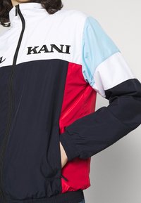 Väriblokkeihin jaettu tuulitakki laivastonsinisellä, punaisella, valkoisella ja vaaleansinisellä. Siinä on vetoketjullinen suljenta, joustavat hihansuut ja rinnan kohdalla sana "KANI".