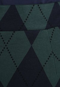 Zelená a námořnická modrá látka se vzorem diamantových tvarů a černými tečkovanými akcenty, s hladkým povrchem a pružným materiálem.