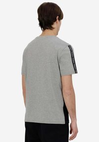 Camiseta de algodón gris con mangas cortas, presenta paneles laterales negros y cinta de marca a lo largo de los hombros. Cuello redondo, ajuste casual.