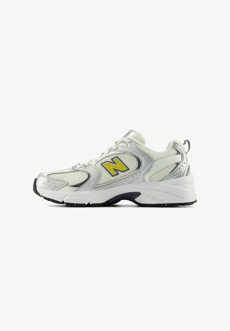 Chaussure de sport blanche et argentée avec des panneaux en mesh, logo "N" jaune sur le côté, semelle noire et blanche, et languette de traction au talon.