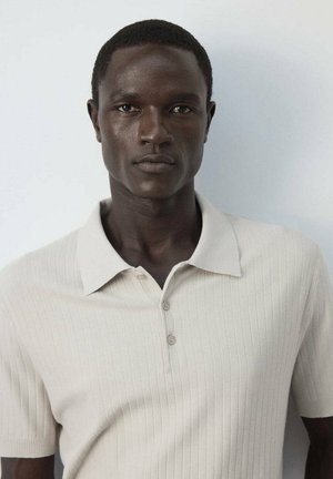 Jeune homme à la peau foncée portant un polo en maille beige clair à manches courtes, debout devant un fond clair uni.