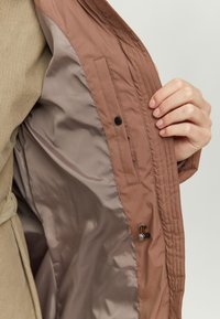 A brown outer jacket with a smooth, light interior. Features a button, an interior pocket, and stitched detailing along the edges.