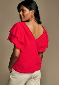 Blusa rossa con maniche a strati fluide, scollatura a V sul retro e orlo ondulato. Realizzata in tessuto testurizzato, abbinata a pantaloni chiari.