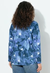 Langärmliges T-Shirt mit Batikmuster in Blautönen und Sternenstrahl-Design; weicher Stoff, lockere Passform und abgerundeter Saum. Getragen mit hochtaillierten Jeans.