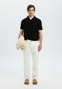 Juodas trumpomis rankovėmis mezginys polo marškiniai, balta marškinėliai, blyškiai balti kelnės, smėlio spalvos rankinė ir rudos spalvos šlepetės, stovintis prieš šviesų foną.