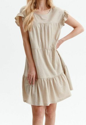 Vrouw met lang blond haar, die een beige knielange jurk met korte mouwen en geplooide lagen draagt, poseert met één hand op haar heup en de andere houdt de zoom van de jurk vast.