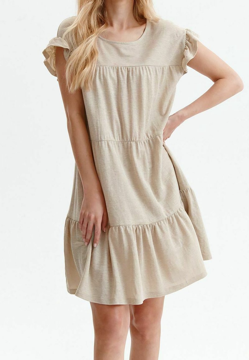 Femme aux longs cheveux blonds portant une robe beige courte avec des manches courtes et des volants froncés, posant avec une main sur la hanche et l'autre tenant l'ourlet de la robe.
