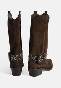 Braune Wildleder-Stiefel bis zum Knie mit einem schwarzen Lederknöchelriemen, der mit runden Metallösen verziert ist, und einem kleinen Blockabsatz.