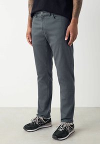 Jeans de mezclilla gris de corte ajustado, con bolsillos delanteros, un pequeño bolsillo para monedas y detalles de costura sutiles. Combinados con zapatillas deportivas negras.