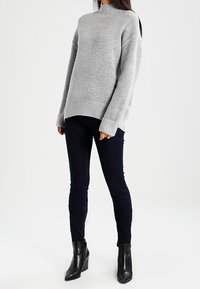 Suéter gris de cuello alto con mangas largas, combinado con jeans oscuros ajustados y botas negras hasta el tobillo. Textura suave, diseño relajado, minimalismo en los detalles.