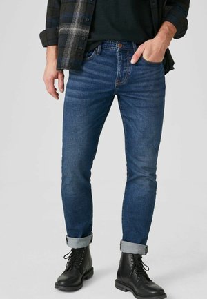 Mann trägt dunkelblaue, hochgekrempelte Slim-Fit-Jeans, schwarze Schnürstiefel, ein schwarzes Hemd und eine dunkel karierte Jacke mit teilweise hochgekrempelten Ärmeln.