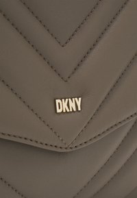 Hnědošedý prošívaný kožený povrch s chevronovým švem a malým zlatým logem DKNY umístěným uprostřed klopy tašky.