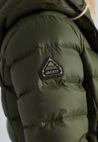 Olijfgroene gewatteerde jas met een quilted textuur, voorzien van een driehoekige zwarte patch met het label "JACK1T" op de linkermouw.