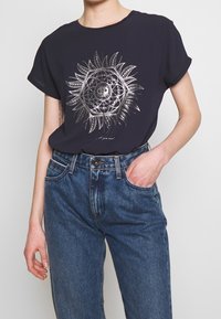 Camiseta de manga corta azul marino con un gráfico metálico del sol con detalles intrincados. Combinada con unos vaqueros de mezclilla azul de cintura alta.