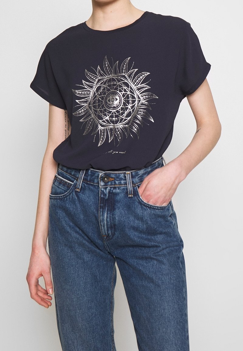 Camiseta de manga corta azul marino con un gráfico metálico del sol con detalles intrincados. Combinada con unos vaqueros de mezclilla azul de cintura alta.