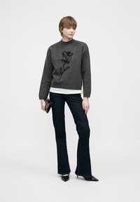 Grijs sweater met zwarte rozenborduursel, gecombineerd met donkere flare jeans. Model houdt een kleine handtas vast en staat tegen een effen achtergrond.
