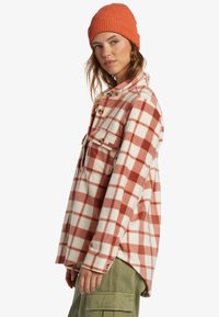Plaid flannel shirt in red and cream with a relaxed fit, two chest pockets, and wooden buttons. Paired with a ribbed orange beanie.