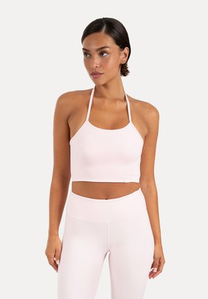 Kobieta w jasnoróżowym topie typu halter i legginsach z wysokim stanem, stojąca w zrelaksowanej pozie na jednolitym tle.