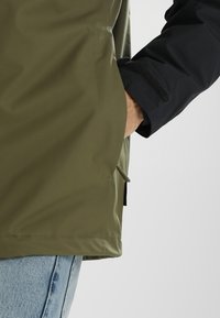 Personne portant une veste verte et noire avec la main dans la poche latérale, jean bleu visible sur un fond clair uni.