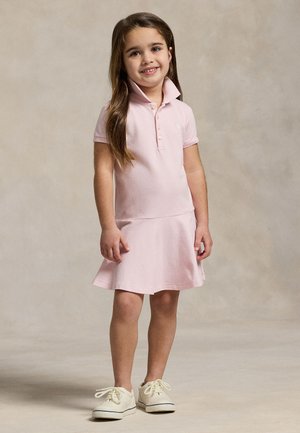 Jeune fille souriante, debout, portant une robe polo rose clair et des baskets blanches sur un fond neutre uni.