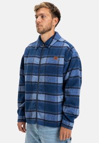 Blue fleece jacket with a check pattern in varying shades of blue and dark lines, featuring a zip closure and a small logo on the chest.
