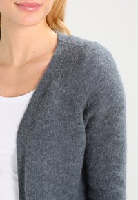 Grå ullkofta med v-ringning, mjuk textur och lätt fluffig finish, kombinerad med en vit skjorta under.