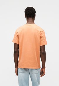 Πορτοκαλί κοντομάνικη μπλούζα από βαμβάκι, με στρογγυλή λαιμόκοψη και άνετη εφαρμογή. Η πλάτη είναι μονόχρωμη με λεία υφή.