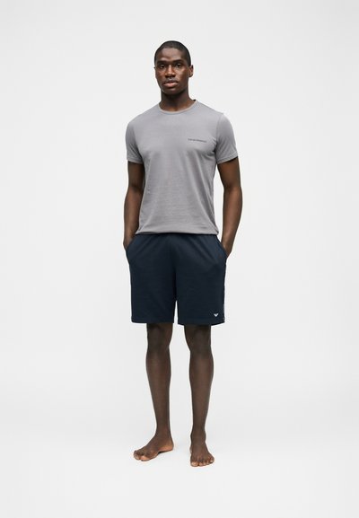 Άνδρας που στέκεται ξυπόλυτος με τα χέρια στις τσέπες, φορώντας γκρι κοντομάνικη μπλούζα και σκούρο μπλε σορτς, μπροστά σε απλό φόντο.