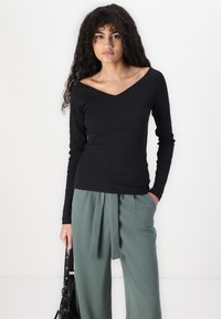 Black ribbed long-sleeve top with a V-neck design, paired with high-waisted green trousers featuring a tied waistband detail.
