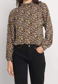 Blusa floreale marrone con un collo alto arricciato, maniche lunghe e una vestibilità ampia, abbinata a jeans neri attillati. Presenta piccoli fiori bianchi e gialli.