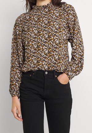 Bruine bloemenblouse met een hoge gerimpelde halslijn, lange mouwen en een losse pasvorm, gecombineerd met zwarte strakke jeans. Heeft kleine witte en gele bloemen.