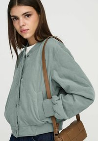 Veste bomber matelassée vert clair avec un col rond, des boutons-pression, des poignets élastiques et une poche latérale, associée à un sac bandoulière marron.