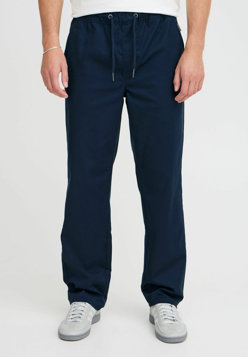 Mørkeblå bukser laget av et glatt stoff, med elastisk midje og snor, samt sidelommer. Kombinert med lysegrå joggesko.