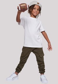 Child wearing a white cotton t-shirt, olive green cargo pants with cuffed ankles, and white sneakers, holding a brown football.