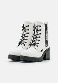 Vita anklarstövlar med svarta snören och dragkedja på sidan, med en kraftig svart klack och strukturerad gummisula. Varumärkesetikett på sidan.