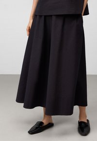Svart texturerad midikjol med veck och avslappnad passform, tillsammans med blanka svarta slip-on skor. Platt, slät yta i en minimalistisk miljö.