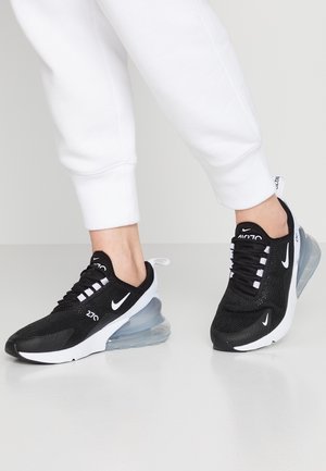 Person iført sorte og hvide Nike Air Max 270 sneakers med gennemsigtige luftpolstringer under sålen og hvide bukser.