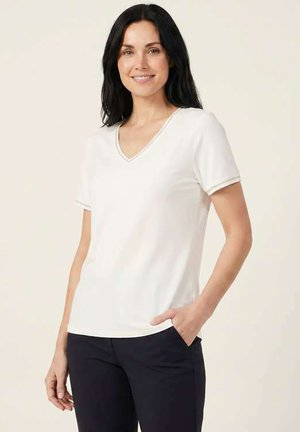 Femme aux longs cheveux foncés portant un haut blanc à manches courtes en V et un pantalon noir, souriante avec une main dans sa poche.