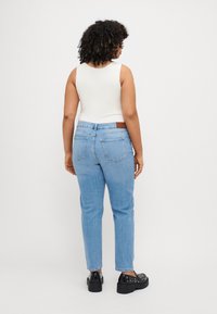 Helle blaue Jeans mit geradem Bein, mit einem braunen Lederpatch am hinteren Bund und an den Taschen. Kombiniert mit einem weißen ärmellosen Oberteil und schwarzen Plateauschuhen.