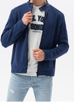 Mann trägt hellblaue, zerrissene Jeans, weißes T-Shirt mit Aufdruck und teilweise geschlossene, marineblaue, strukturierte Jacke und richtet den Reißverschluss.