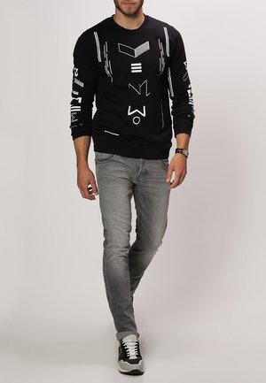 Mężczyzna idzie ubrany w czarną bluzę z białymi abstrakcyjnymi symbolami, szare dopasowane jeansy, czarno-białe sneakersy oraz zegarek na rękę.